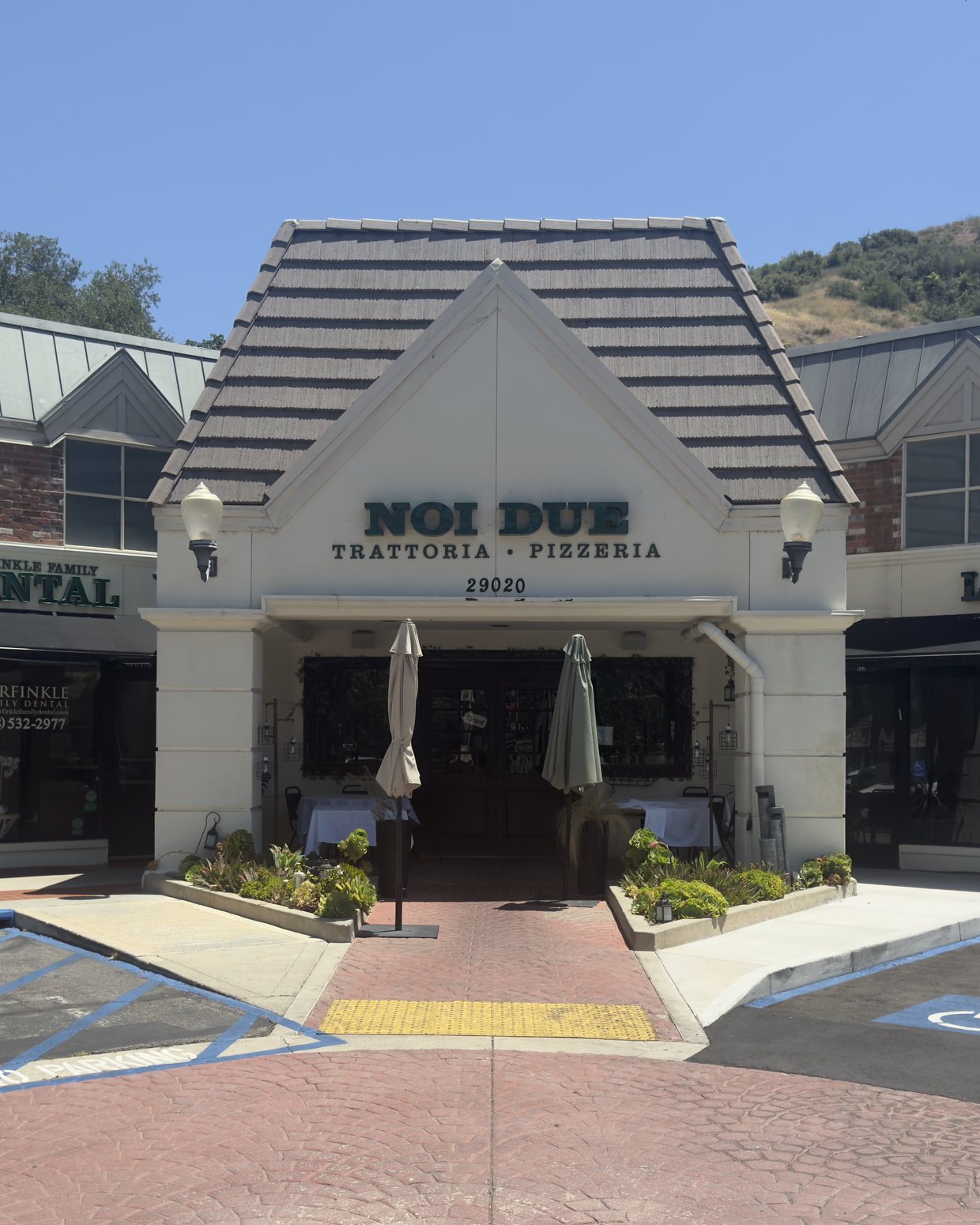 The gabled entrance to Noi Due Trattoria at Agoura Village Center, flanked by Garfinkle Family Dental and L&R Salon, with the Santa Monica Mountains behind